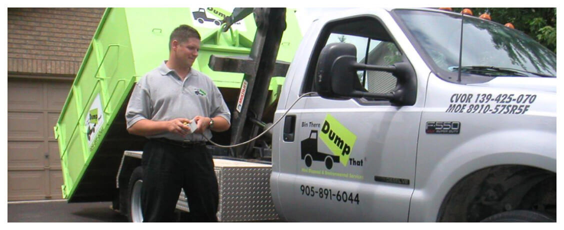 A man stands next to a white truck with a green dumpster, holding a remote control. The truck's doors display company information and a logo that reads "Bin There Dump That.